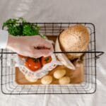 Dłoń trzyma kosz z pieczywem, ziemniakami, pomidorami i zieleniną